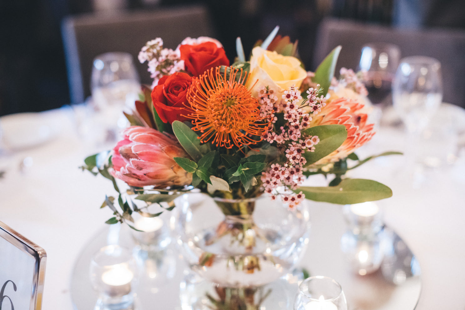 Elegant wedding table centerpiece featuring roses, proteas, and delicate fillers in a glass vase.