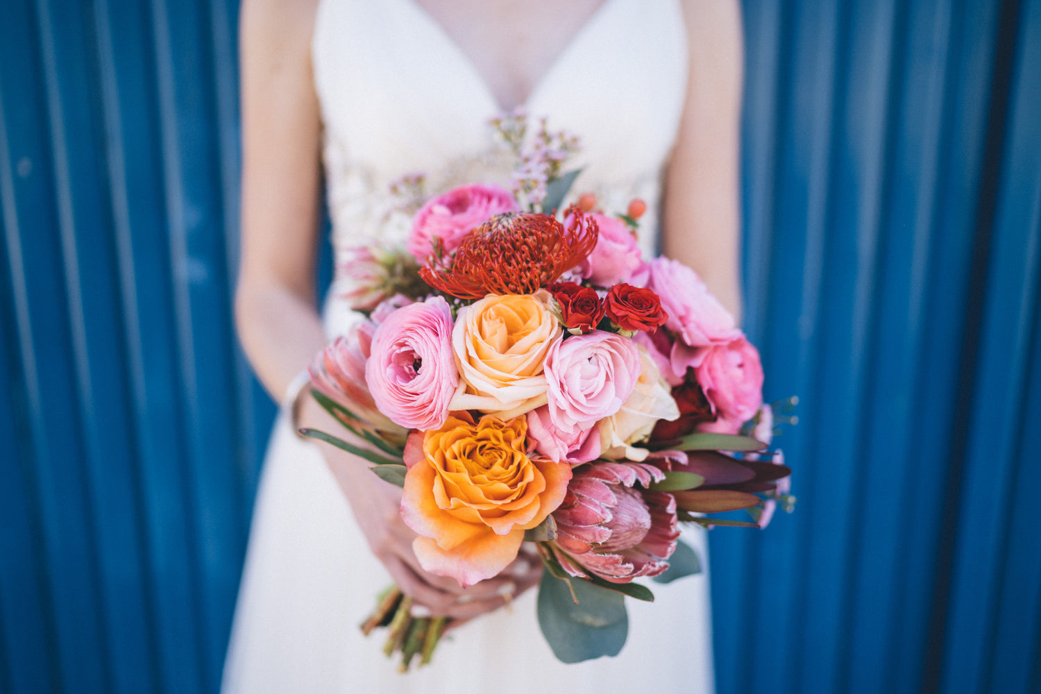 Bride holding a colorful bouquet with pink, orange, and red flowers against a blue background.