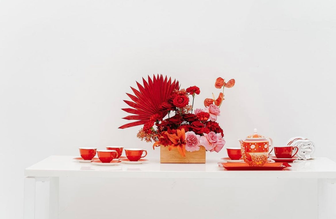 Modern table setting featuring a vibrant floral arrangement with red and pink flowers and red tea set on a white table.