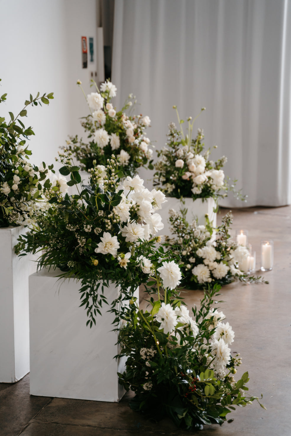 Elegant white floral arrangements on white podiums in a ceremony hall with soft lighting and candles.