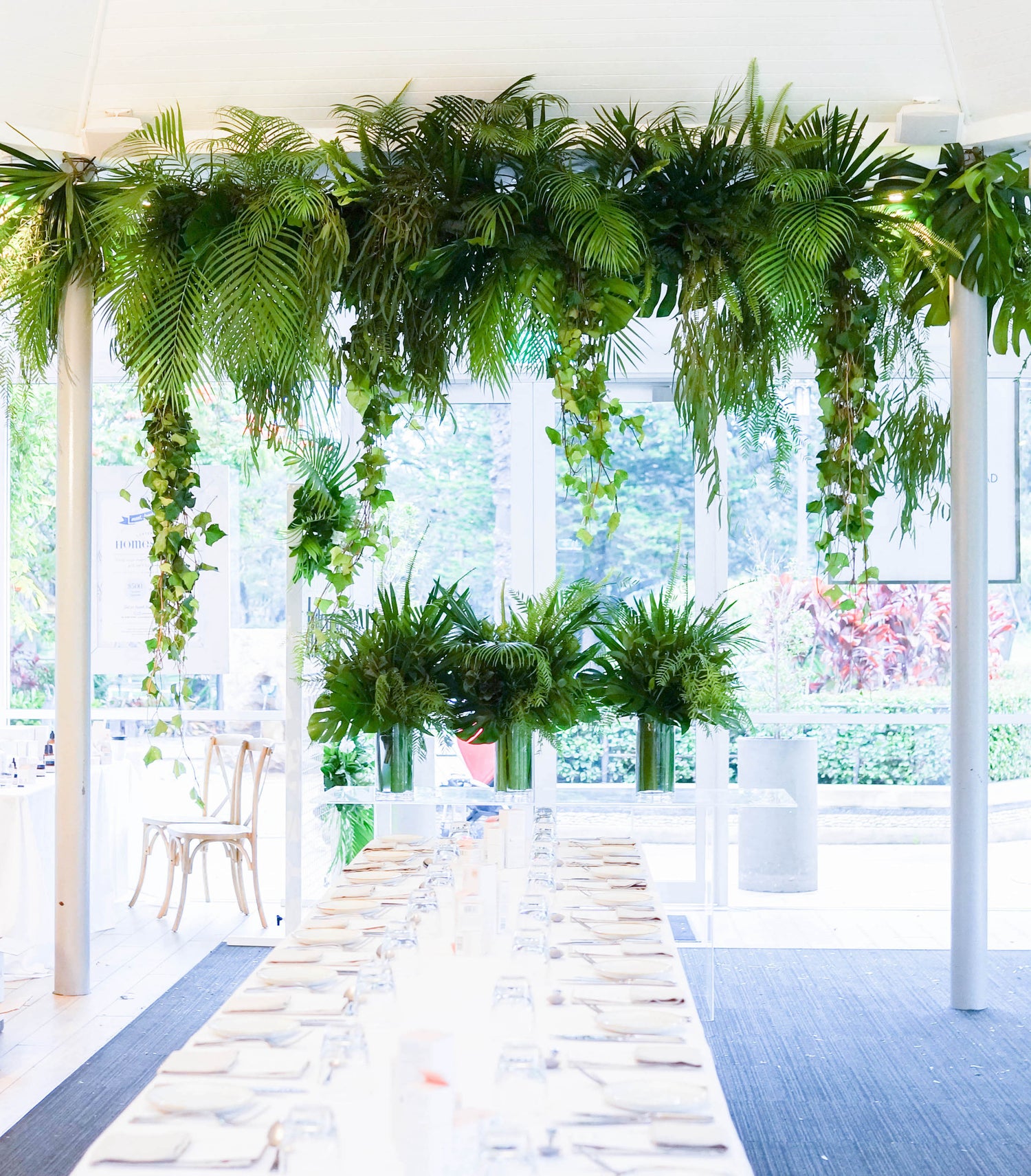 legant dining setup with a long table, white tablecloths, and lush green hanging plants and centerpieces in a bright, glass-walled venue.