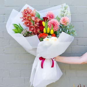 Bouquet of vibrant spring flowers wrapped in white paper held against a brick wall.