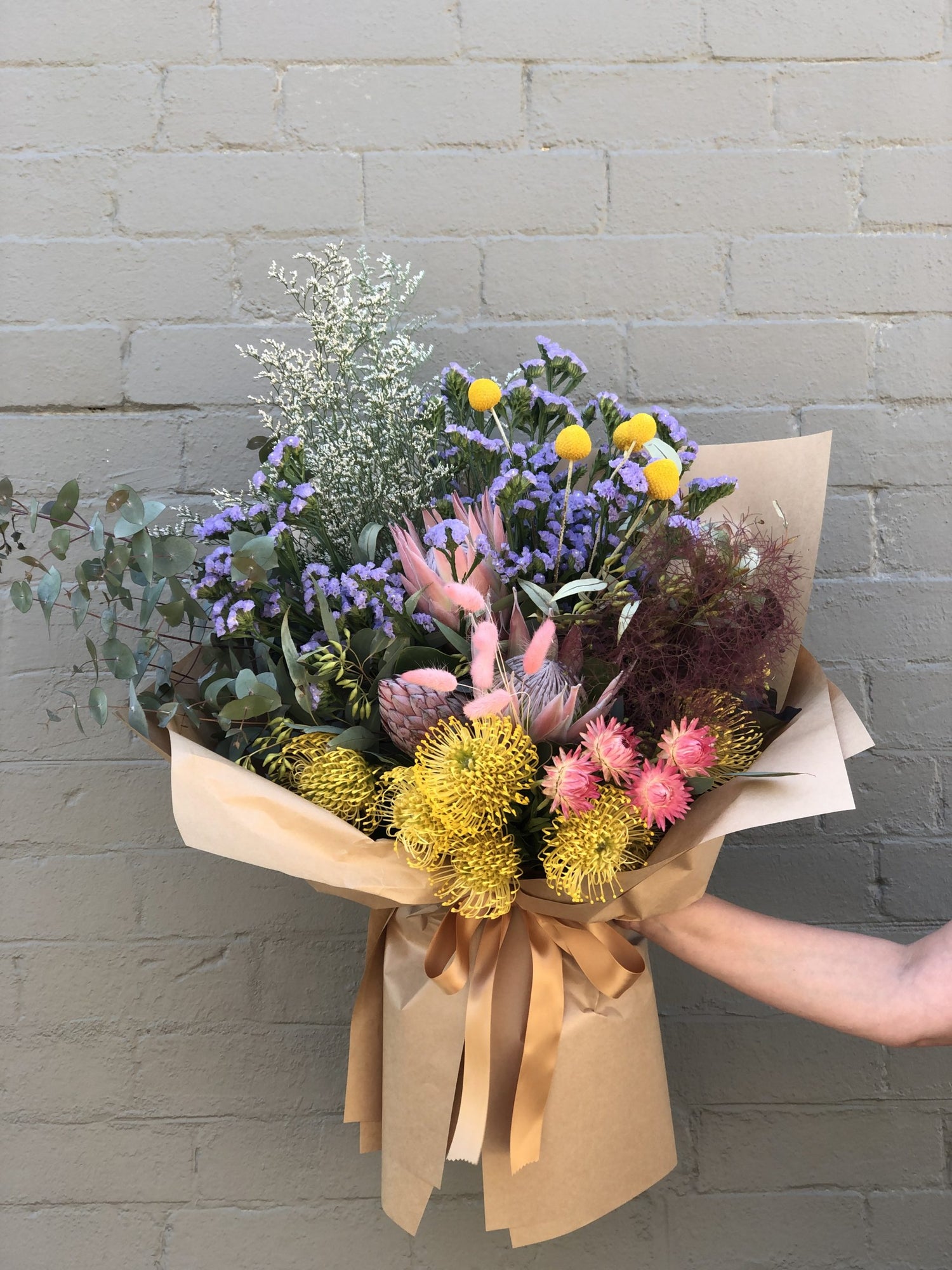 Hand-held bouquet featuring exotic pink and yellow flowers with rustic brown paper wrapping.