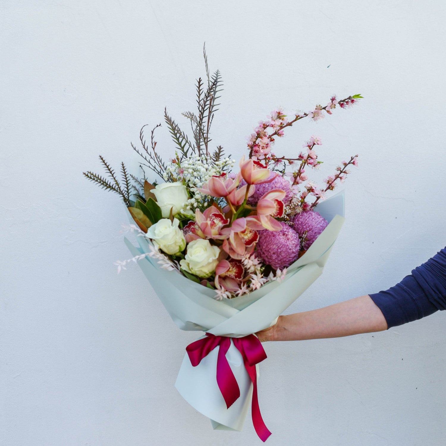 Elegant floral bouquet in pastel shades with roses and branches, wrapped in light blue paper and pink ribbon.