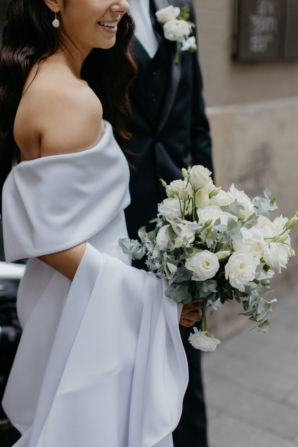 Bride in an off-shoulder gown holding a white floral bouquet with greenery.