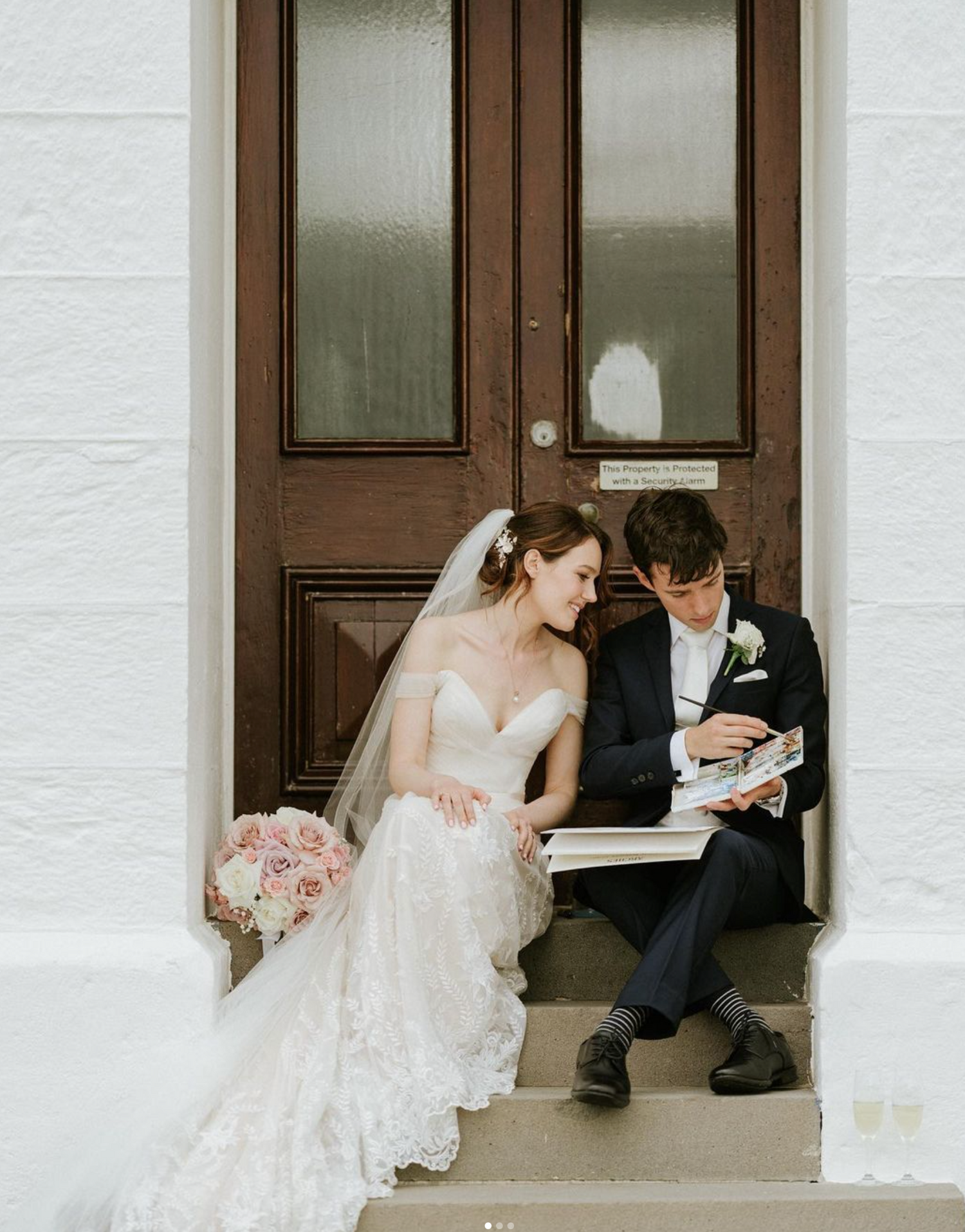 Bride and groom sitting on steps, sharing a moment with a bouquet of pink roses beside them.