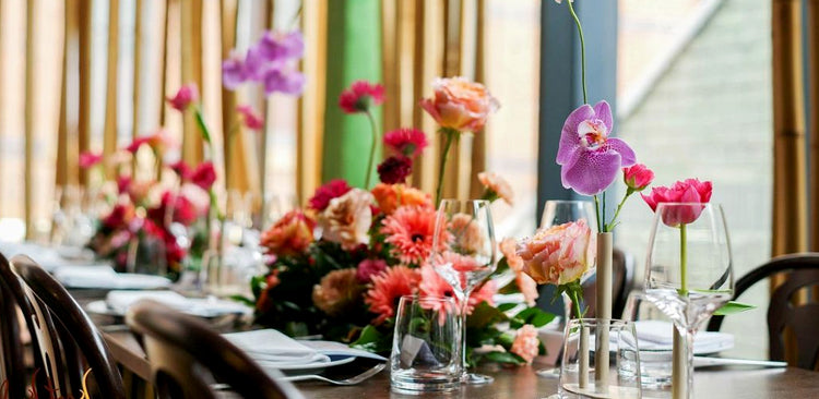 Long dining table adorned with vibrant floral centerpieces featuring orchids, roses, and daisies in a modern restaurant setting.