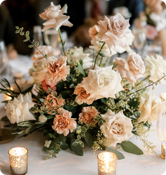 Elegant floral centerpiece with creamy roses and lush greenery on a wedding reception table lit by candlelight.