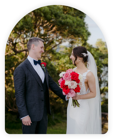 Bride and groom smiling at each other, holding a bouquet of pink and red roses in a natural setting.