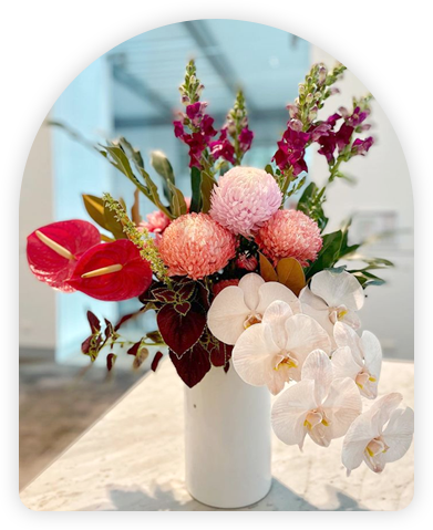 Vibrant and colorful floral arrangement in a white vase, featuring pink pompoms, red anthuriums, and white orchids.