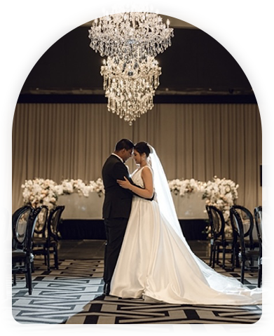 Newlyweds sharing a romantic moment under a grand chandelier in a luxurious ballroom with floral decorations in the background.