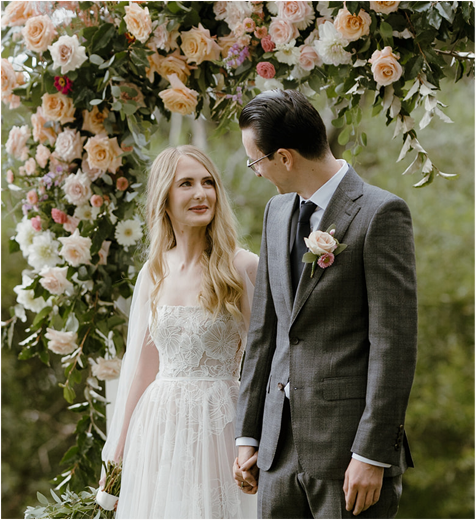 Bride and groom holding hands under a flower arch.