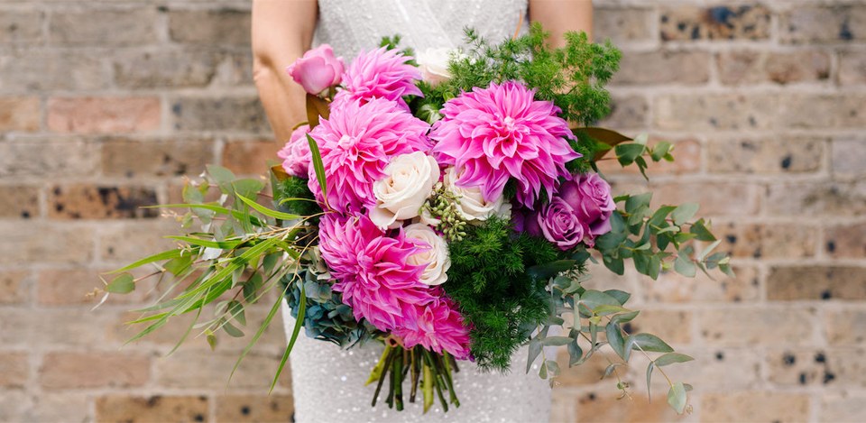 Bride holding a large bouquet of pink, white, and purple flowers in front of a brick wall