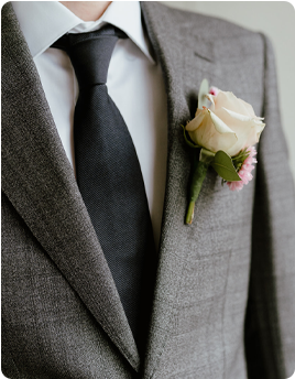 A groom in a gray suit with a black tie and a white rose boutonniere.