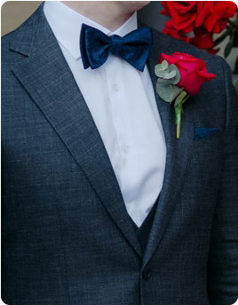 A groom in a dark suit with a red rose boutonniere and a blue bow tie.